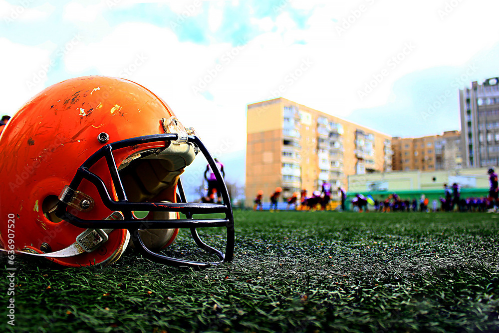 A large orange american football helmet stands close up on the green grass of the stadium A large orange american football helmet stands close up on the green grass of the stadium