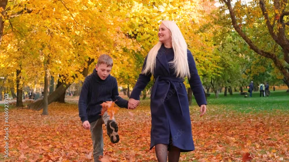 Mother and son are walking in the park, holding hands and tossing autumn leaves up with their feet. Horizontal slow motion video Mother and son are walking in the park, holding hands and tossing autumn leaves up with their feet. Horizontal slow motion video