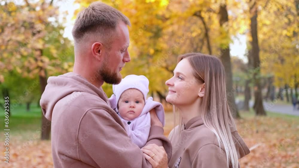 Young mother and father are walking with a newborn baby in the autumn park. Parents in identical tracksuits smile and look at the child. Horizontal slow motion video Young mother and father are walking with a newborn baby in the autumn park. Parents in identical tracksuits smile and look at the child. Horizontal slow motion video
