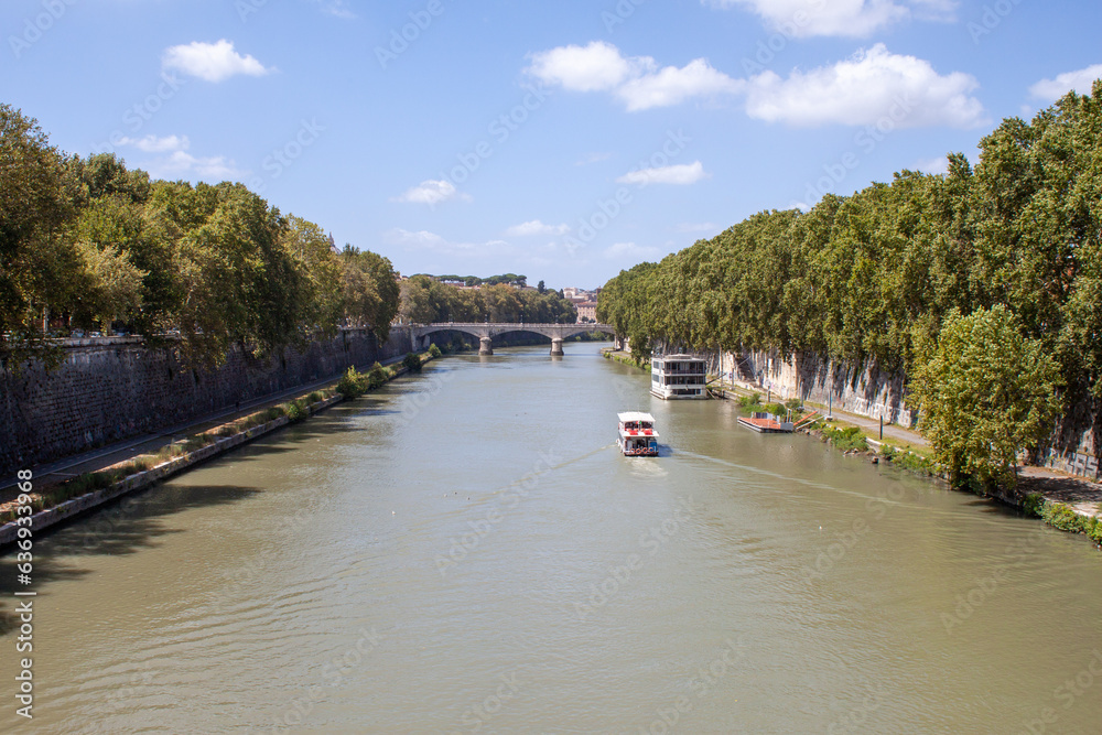 Fototapeta premium Rome, Italy - August 06, 2023: View of the Tiber from Ponte Sisto on a sunny day, cloudy water and a boat going up the river.