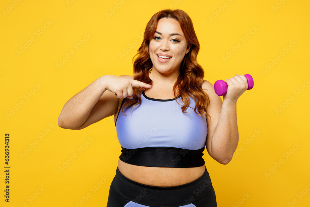 Young chubby overweight plus size big fat fit woman wear blue top warm up training hold show point index finger on dumbbell isolated on plain yellow background studio home gym. Workout sport concept. Young chubby overweight plus size big fat fit woman wear blue top warm up training hold show point index finger on dumbbell isolated on plain yellow background studio home gym. Workout sport concept.