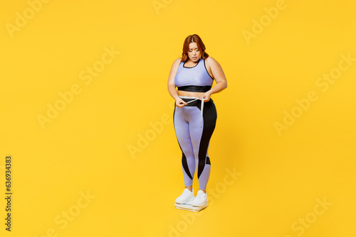 Full body sad young plus size big fat fit woman wear blue top warm up train stand on scales check result look at measure tape isolated on plain yellow background studio home gym Workout sport concept.