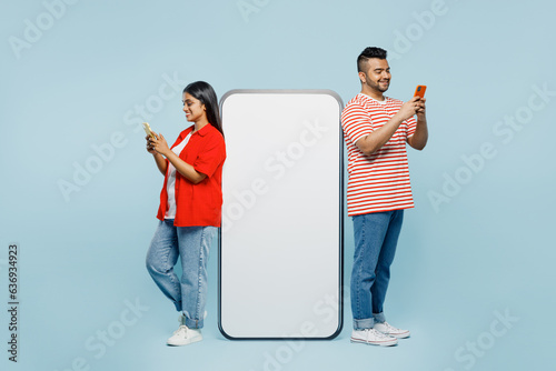 Full body young couple two friends family Indian man woman wearing red clothes t-shirts together big huge blank screen area mobile cell phone using smartphone device isolated on plain blue background.