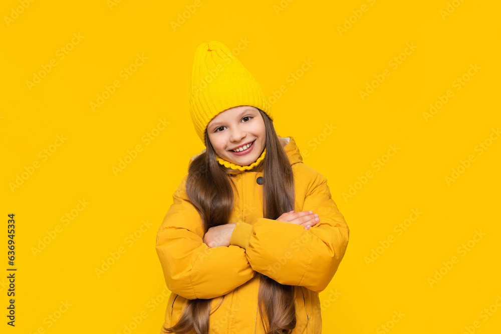 A young girl on an autumn day. A child in a beanie hat and a warm down jacket. A charming little lady is happy about the coming autumn. Fashionable clothes for teenagers. Yellow isolated background. A young girl on an autumn day. A child in a beanie hat and a warm down jacket. A charming little lady is happy about the coming autumn. Fashionable clothes for teenagers. Yellow isolated background.