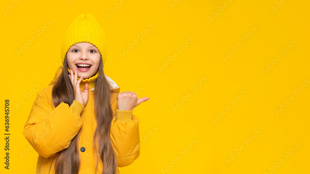 A happy little girl in an autumn jacket and hat points to your advertisement and smiles broadly. Yellow isolated background. Copy space. Banner.