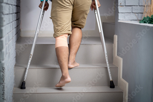Fotografia Closeup leg young asian man walking on stair with crutch at home, close-up man walking while bandage with leg, rear view, medical and lifestyle concept