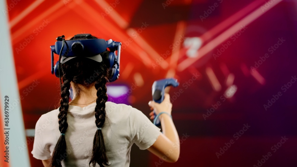 The child use tracking controllers in her hands, moves actively immersing herself in modern game. The little girl wearing VR headset plays video game. Behind girl there is screen with blur background The child use tracking controllers in her hands, moves actively immersing herself in modern game. The little girl wearing VR headset plays video game. Behind girl there is screen with blur background