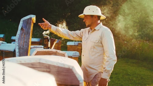 Beekeeper using special equipment. Apiarist man smoking honeycomb of beehive using hive smoker in apiary. Beekeeper working in apiary on sunny day. Honey bees, beekeeping, honeycraft. photo