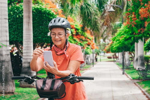 Smiling senior cyclist woman with helmet riding electric bike in the city park stops to answer the phone. Authentic retirement living and healthy lifestyle concept.