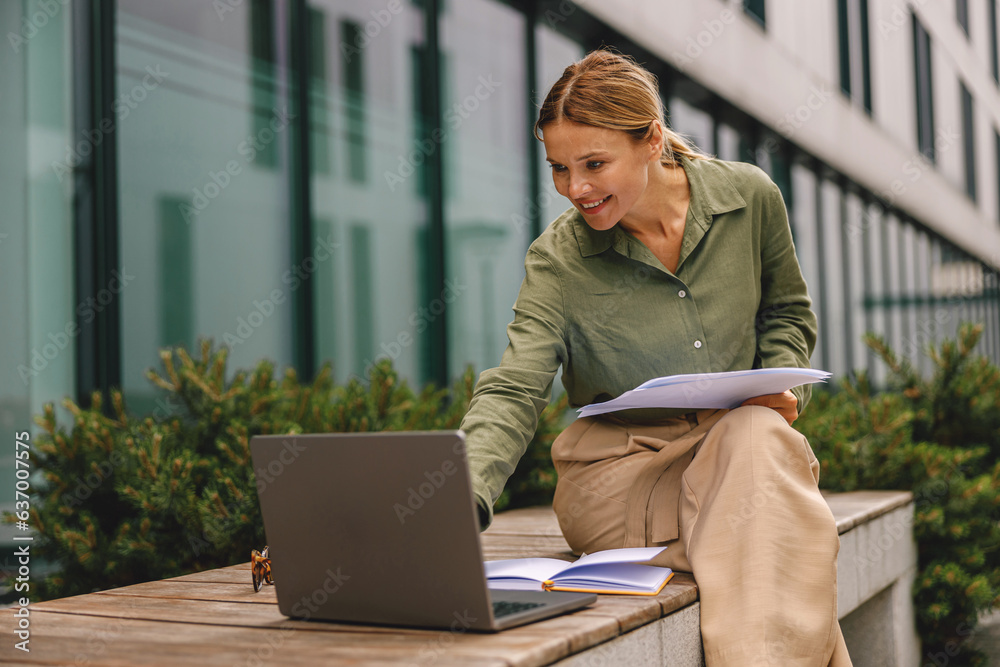 Woman analyst working with documents and use laptop sitting outside on modern building background - obrazy, fototapety, plakaty 