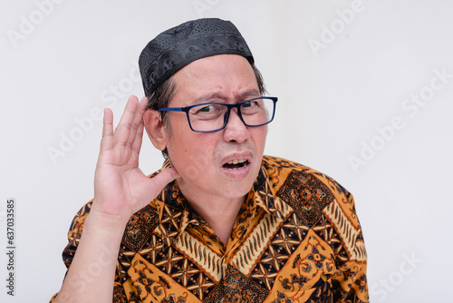 A deaf indonesian man asks someone to speak louder. Gesturing with hand on ear. Wearing a batik shirt and songkok skull cap. Isolated on a white background.
