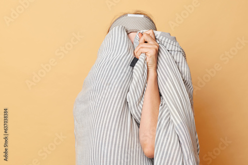 Unknown unrecognizable woman wrapped blanket and wearing sleeping eye mask isolated over beige background hiding her face do not want to wake up. photo