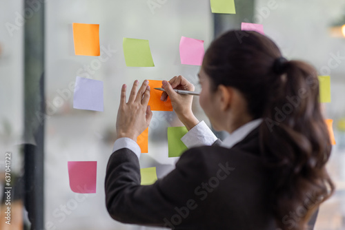 Young Serious Asian Creative team use post it notes to share idea sticky note on glass wall. Asian business people design planning and Brainstorming thinking sticky History notes concept.