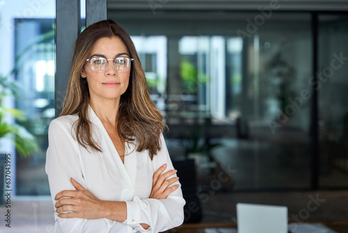 Confident 45 year old Latin professional mid aged business woman corporate leader, mature female executive manager wearing eyeglasses standing in office arms crossed looking at camera, portrait.