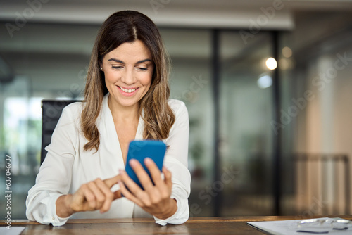 Smiling happy mature mid aged 40 years old professional business woman executive holding smartphone using cell phone mobile apps looking at cellphone working on tech device sitting at office desk. photo