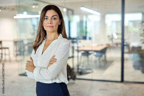 Confident mid aged Latin professional business woman corporate leader, happy mature female executive, elegant lady manager of middle age standing in office arms crossed looking at camera, portrait.