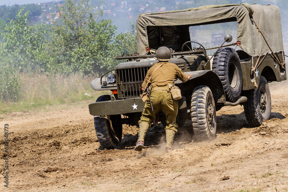 Fototapeta premium Historical reconstruction. An American infantry soldier from the World War II prepare to attack hidden behind a milirary vehicle between smoke and dust. View from the back.