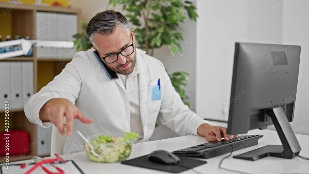 Grey-haired man doctor eating salad talking on smartphone at the clinic Grey-haired man doctor eating salad talking on smartphone at the clinic
