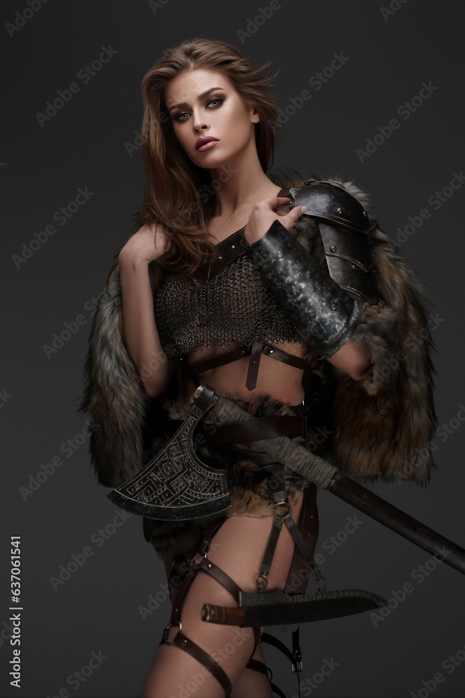 Stunning Viking model dressed in chainmail armor and fur, posing with an axe against a gray wall
