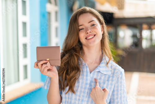 Young redhead woman holding a wallet at outdoors with surprise facial expression