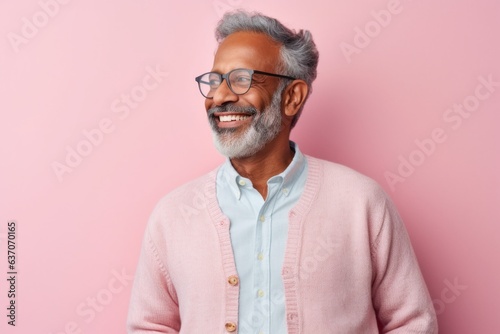 Portrait of happy mature Indian man in eyeglasses smiling at camera while standing against pink background