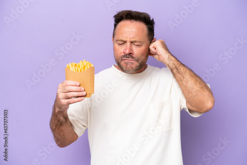 Middle age caucasian man holding fried chips isolated on purple bakcground frustrated and covering ears © luismolinero