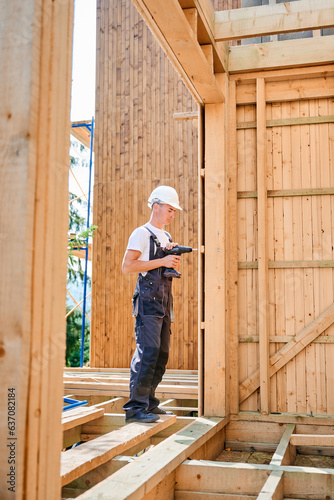Carpenter constructing wooden framed house. Man worker cladding facade of house, using a screwdriver, wearing work overalls and helmet. Concept of modern eco-friendly construction.