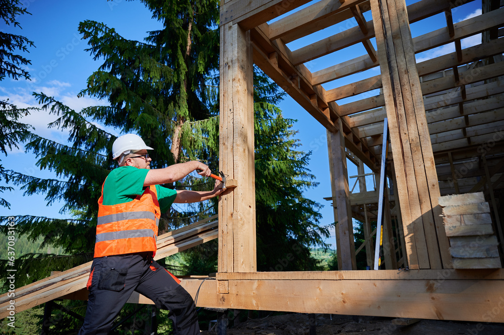 Carpenter constructing two-story wooden frame house near the forest. Man hammering nails into structure while wearing protective helmet and construction vest. Concept of modern ecological construction Carpenter constructing two-story wooden frame house near the forest. Man hammering nails into structure while wearing protective helmet and construction vest. Concept of modern ecological construction