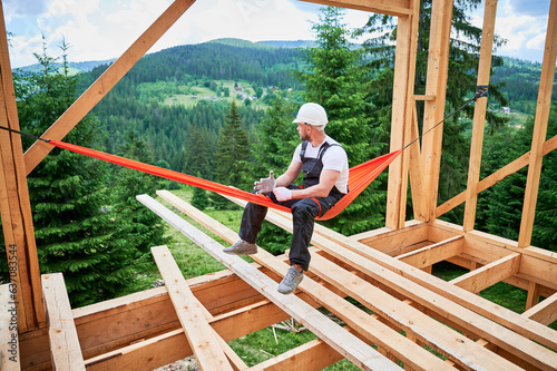 Carpenter, working on construction of wooden-framed house, takes break in hammock, dressed in protective helmet, work overalls and gloves. He enjoys picturesque view of mountains and forests.