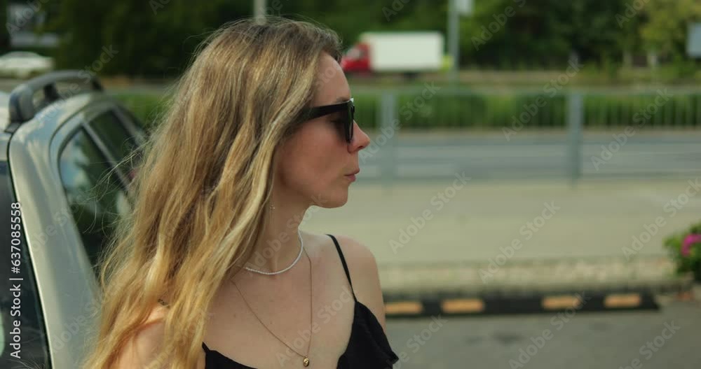 Young woman with long hair shake head and put on sunglasses on sunny summer day. Side view of beautiful confident and contented female driver in black dress stand outdoors at car on parking lot. Young woman with long hair shake head and put on sunglasses on sunny summer day. Side view of beautiful confident and contented female driver in black dress stand outdoors at car on parking lot.