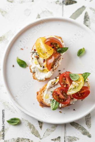 Bruschetta sandwiches with tomatoes, cream cheese, olive oil and basil on white plate on printed tile background close up, top view. Traditional italian antipasti
