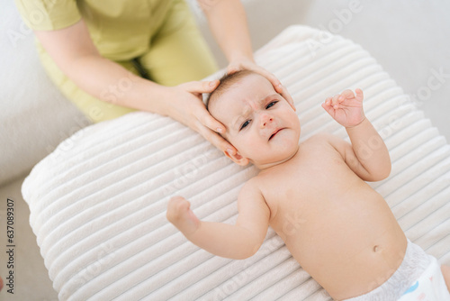 Top view of sad infant baby being manipulated by unrecognizable osteopathic manual therapist or physician. Close up of female masseuse massage head of small newborn kid. Concept of infant healthcare.