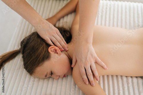 Top view of cute little girl having neck, shoulder and back massage by unrecognizable female masseuse lying on massage table. Adorable preteen kid getting physiotherapy from masseuse for body care.