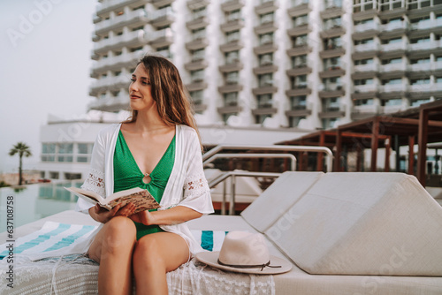 Chica joven en hamaca con smartphone tomando fotografías y leyendo en piscina de hotel de lujo photo