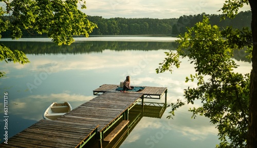 Bilde på lerret Young female sitting on a wooden dock at a lake, surrounded by tall trees and fo