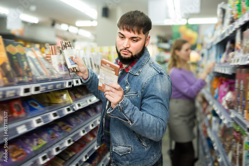 Принти на полотні Young bearded guy choosing chocolate on shelves in grocery store