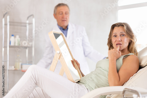 Unsatisfied middle-aged woman patient lying on clinical chair holding mirror and doctor looking with bewilderment