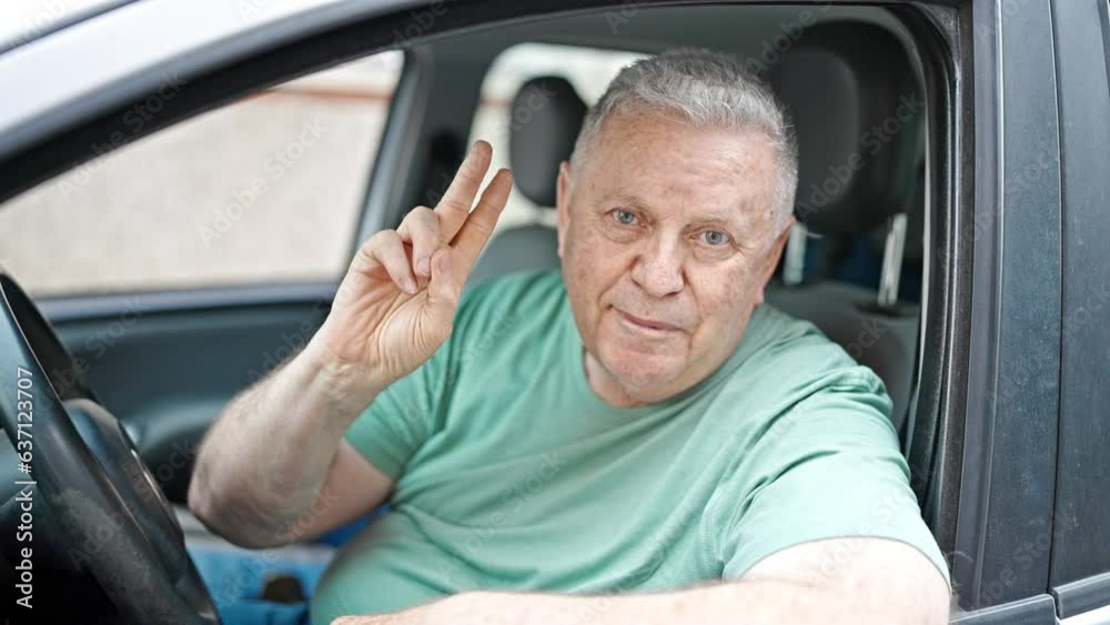 Middle age grey-haired man smiling confident sitting on car doing victory gesture at street