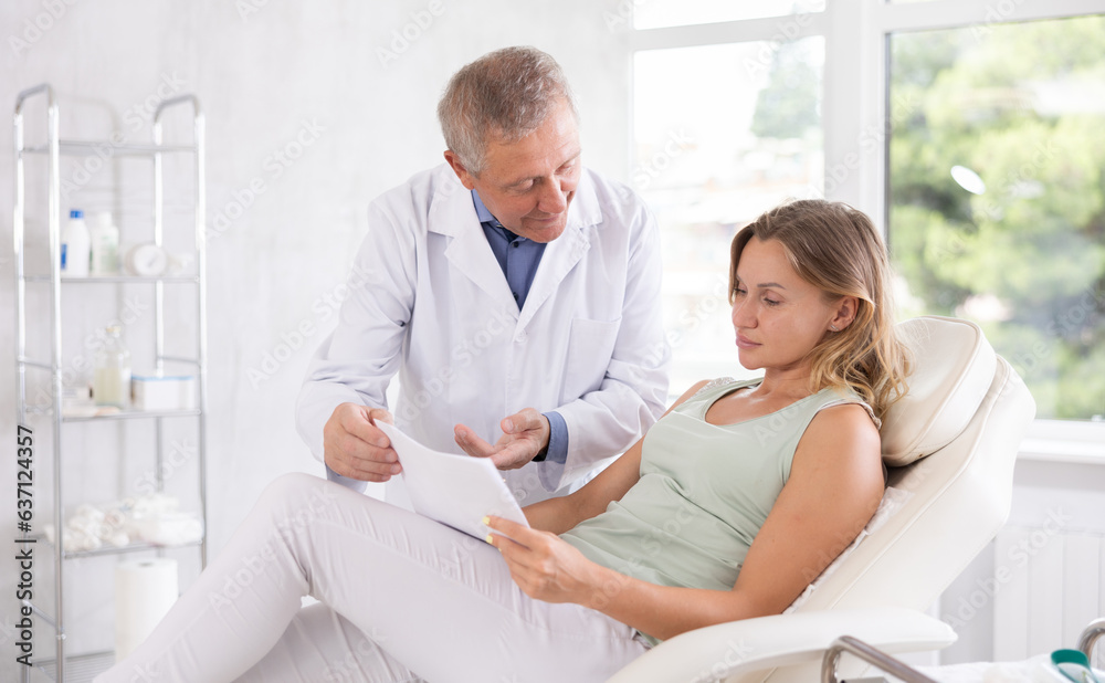 Obraz premium Senior man doctor gives middle-aged female patient sitting in chair medical documents for review before carrying out cosmetic procedure. Medical documentation, questionnaire, patient survey