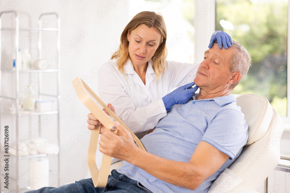 Obraz premium Mature male patient of cosmetology office sitting in chair with mirror in hands and discussing with female doctor need for rejuvenation procedure 