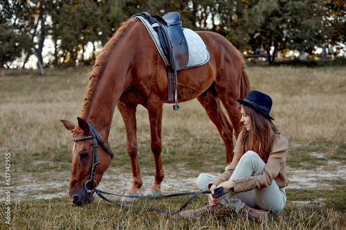 In the countryside, a young woman in black hat sits on grass near a brown horse grazing grass. Horses being fed. Gift certificate for hippotherapy and emotions. Horse in sunset light in the evening