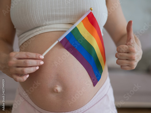 A woman holds a rainbow flag against the background of a naked pregnant belly and shows a thumbs up. 