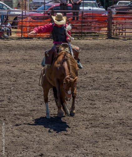 A cowboy is riding a bucking bronco at a rodeo in an arena. The horse has 2 legs off the ground. The cowboy is wearing red with a black hat. There is a red metal gate behind. They are in a dirt arena.