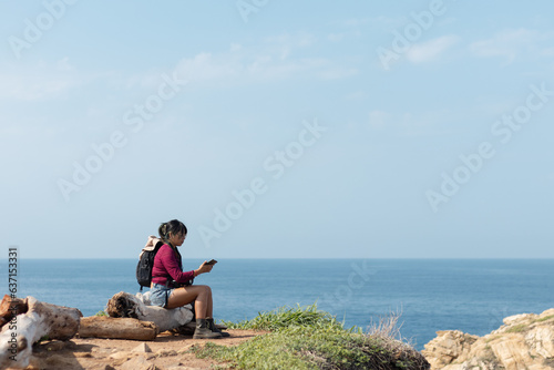 Young tourist sitting on a log checking her phone and texting in the morning near the camping area on the beach of Mazunte Oaxaca © Rangel