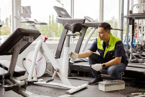 Professional asian male service repair worker or sport fitter using bicycle and treadmill service equipment, supervising and securing fitness equipment in indoor gym. photo