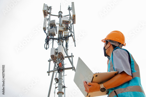 Helmeted asian male engineer works field with telecommunication tower that controls mobile electrical installation using laptop to monitor and maintain 5G networks installed in high rise buildings.
