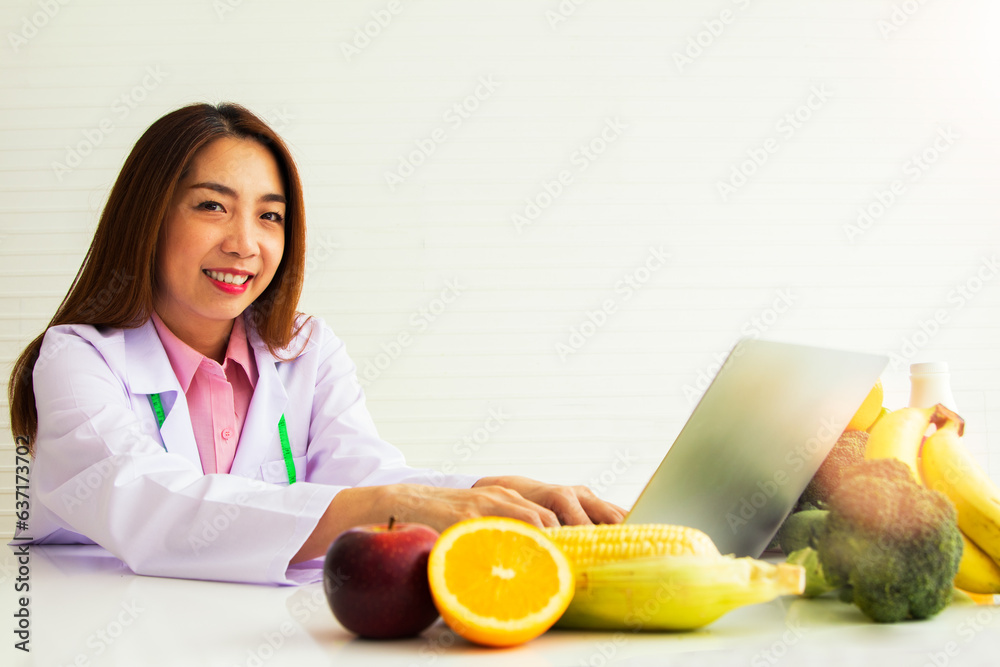 Health care concepts : Professional asian nutritionist, happy wearing white coat on the desk with vegetables and laptops to publish online media suggesting food, vegetables, fruits with nutrition. Health care concepts : Professional asian nutritionist, happy wearing white coat on the desk with vegetables and laptops to publish online media suggesting food, vegetables, fruits with nutrition.