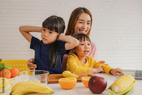 Happy mother teaches two young daughters how eat fruits vegetables healthy nutrition while listening but the eldest daughter is deaf and disinterested covering her ears with frowning and touchy hands. photo