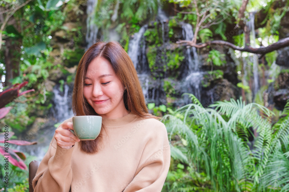 Portrait image of a young woman holding and drinking coffee while sitting in the garden with waterfall Portrait image of a young woman holding and drinking coffee while sitting in the garden with waterfall