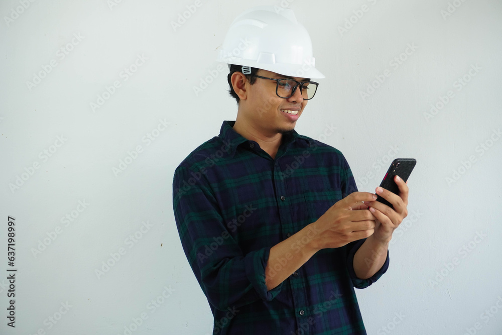 smiling young Asian male engineer wearing white hard hat holding mobile phone for construction work isolated on white background. smiling young Asian male engineer wearing white hard hat holding mobile phone for construction work isolated on white background.